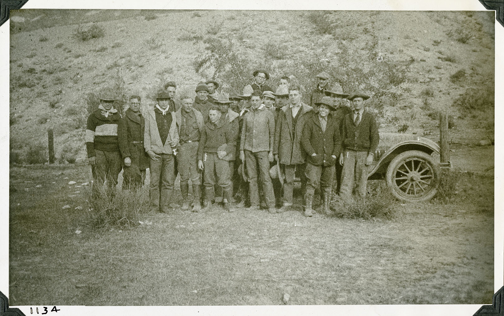 This is an historic black and white photograph from the Scotty's Castle Historic Photograph Collection, Death Valley National Park of twenty-one young men and older men posing in front of a vehicle parked by a fence. Most are college-aged young men. Most have hats, heavy coats or sweaters, jeans, and solid footwear. Steep, barren hillside behind. Number in black ink in lower left corner.