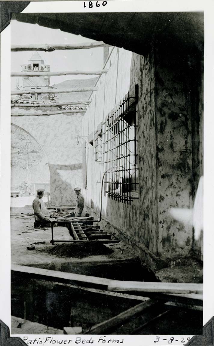 This is an historic black and white photograph from the Scotty's Castle Historic Photograph Collection, Death Valley National Park of two men in white overalls and caps waste deep in trench with wooded forms. Extensive excavation and forms in foreground. Stucco wall with metal grate over window on side. Stucco arch at end. Tower in background. Inscriptions in black ink along lower border.