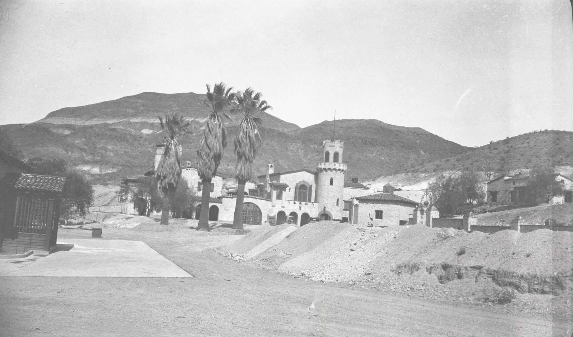 This is an historic black and white photograph from the Scotty's Castle Historic Photograph Collection, Death Valley National Park of a row of piled, excavated dirt. Large, complex, Spanish-style building in background with arches, turrets, and towers. Another structure on left with only covered doorway showing – red tile roof and carved columns on side walls. Area has concrete parking pad. Three large palm trees in center. Desert hillside behind structures.