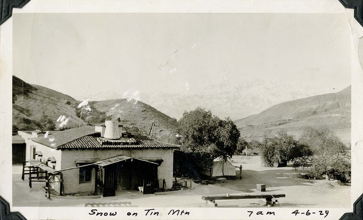 This is an historic black and white photograph from the Scotty's Castle Historic Photograph Collection, Death Valley National Park of white building with red tiled roof in a semi-barren yard and a white wall tent. Overexposed in background in a mountain range with snow on top. Inscriptions in black ink along lower border.
