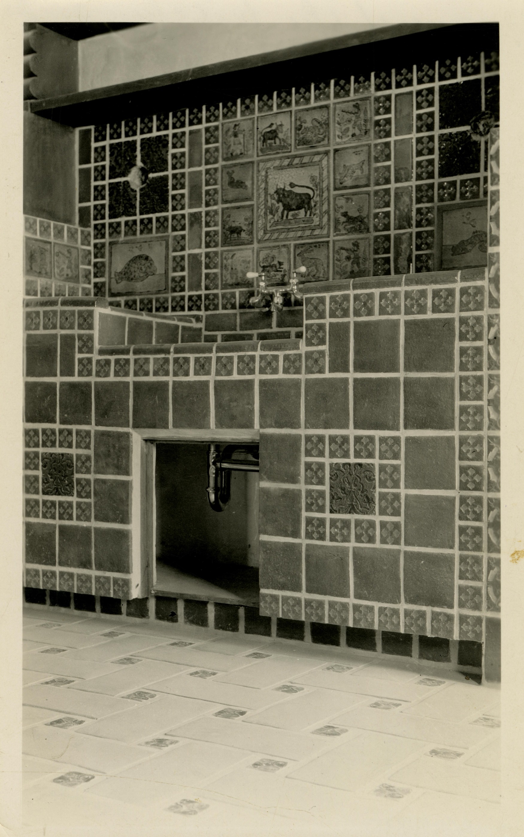 This is an historic black and white photograph from the Scotty's Castle Historic Photograph Collection, Death Valley National Park of kitchen interior with nearly ever surface tile. Tiled kitchen sink and cupboard area. Back splash area behind sink has Spanish bull motif and circular holes for future fixtures. Sink is set down in countertop with two, ivory handle faucets. Sink cupboard below has no door yet revealing pea trap. Floor is tiled.