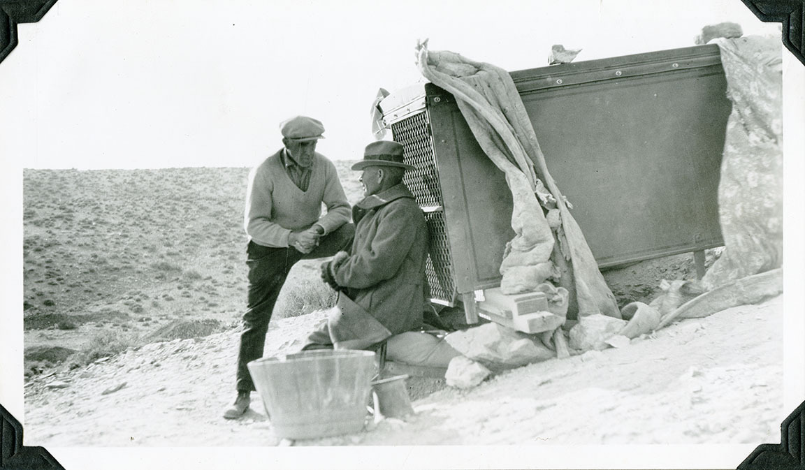 This is an historic black and white photograph from the Scotty's Castle Historic Photograph Collection, Death Valley National Park of one man visiting intently with another man seated in front of a very large metal box. One man is standing but with leg up on box, leaning on it. Other man is seated. Both men dressed for cold weather. Pieces of canvas or quilts secured by rocks on corners of metal box. Large wooden tub and small metal pail in foreground. Sparce, low vegetation on distance hillside.