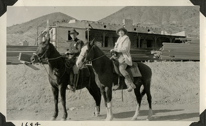 This is an historic black and white photograph from the Scotty's Castle Historic Photograph Collection, Death Valley National Park of Albert M. Johnson and Edna Devlin pose on horseback in front of Scotty's Castle Hacienda. Circa 1928. Photographed by Mat Roy Thompson.