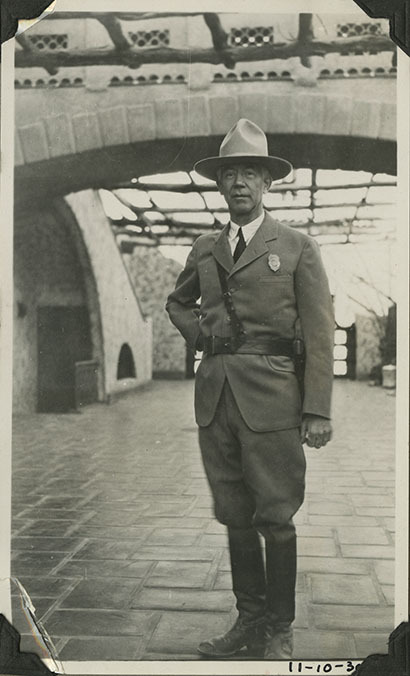 This is an historic black and white photograph from the Scotty's Castle Historic Photograph Collection, Death Valley National Park of Albert M. Johnson wearing sheriff's uniform and standing in Scotty's Castle Annex Enclosed Patio. November 10, 1930. Photographed by Mat Roy Thompson.