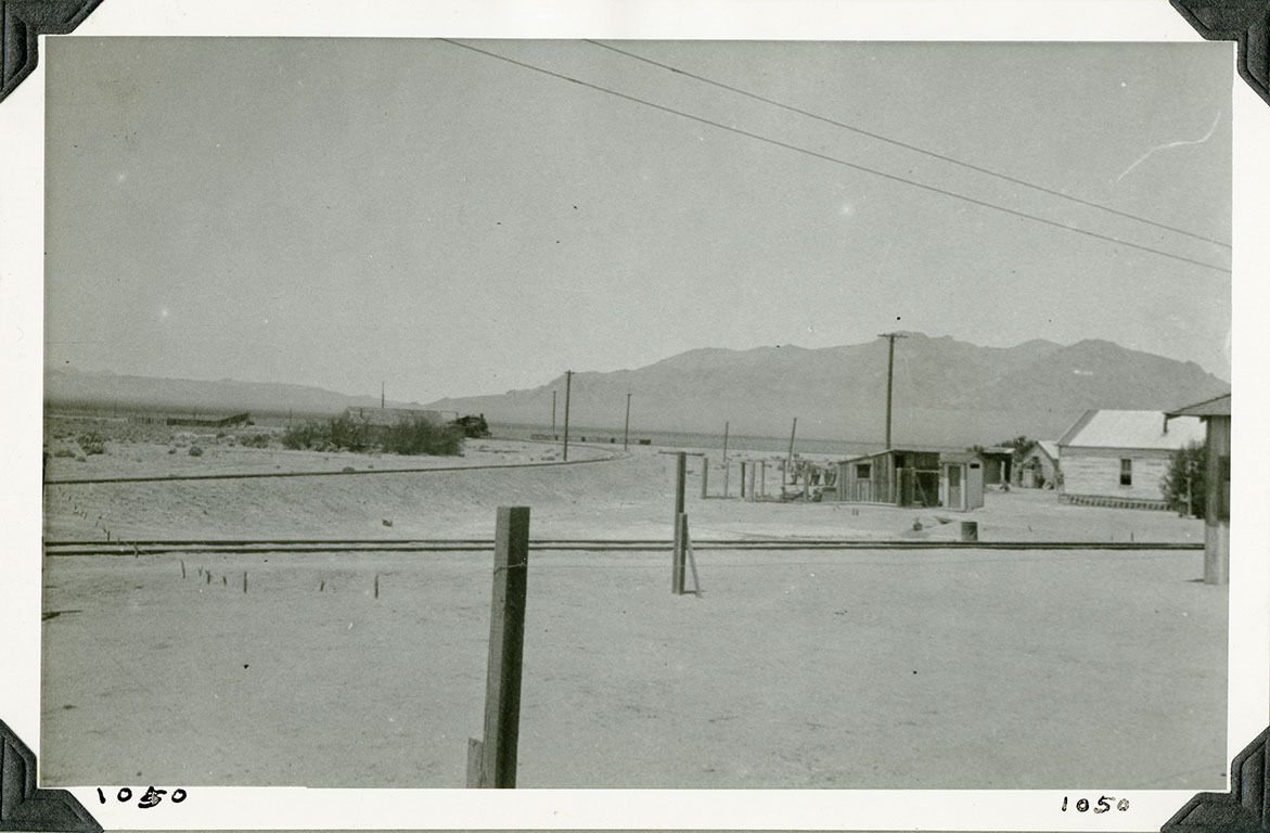 This is an historic black and white photograph from the Scotty's Castle Historic Photograph Collection, Death Valley National Park of two railroad tracks with residential wood buildings on right. One track runs across length of photo. Other track curves off into the distance. Power lines run across upper right corner. Utility poles parallel to tracks. Mountains in distance. Dirt and sand in foreground. Number in black ink in lower left and right corner.