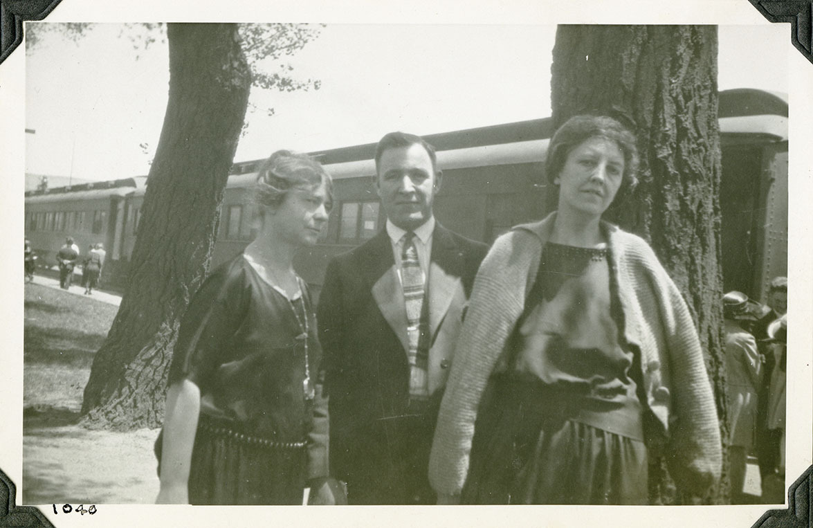 This is an historic black and white photograph from the Scotty's Castle Historic Photograph Collection, Death Valley National Park of three people in front of a passenger train. Woman on left in 1920s style dress and large drop necklace. Man in middle in suite and tie. Woman on right with drop waist dress and light sweater. Large tree trunks between them and train. People mingling along walkway in front of train. Number in black ink in lower left corner.