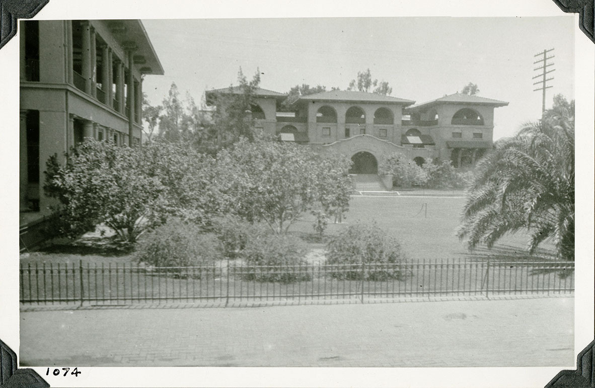This is an historic black and white photograph from the Scotty's Castle Historic Photograph Collection, Death Valley National Park of large Spanish style mansion in large landscape yard. Another large, similar style building on left edge. Wrought iron fence in front. Utility pole on right edge. Number in black ink in lower left corner.
