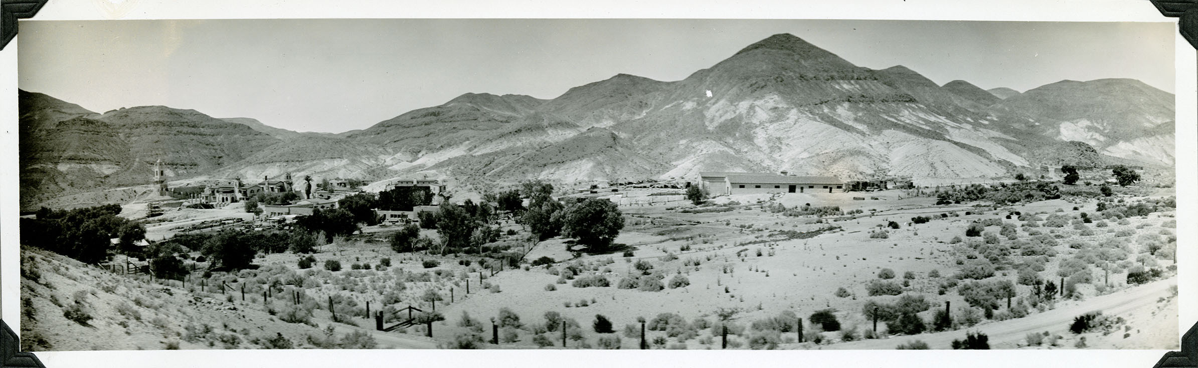 This is an historic black and white panoramic photograph from the Scotty's Castle Historic Photograph Collection, Death Valley National Park of large building and landscape complex in middle of barren, desert scene. Road and fence line in foreground. Middle ground is mostly trees and fields. Then a line of structures at base of barren, desert hillsides.