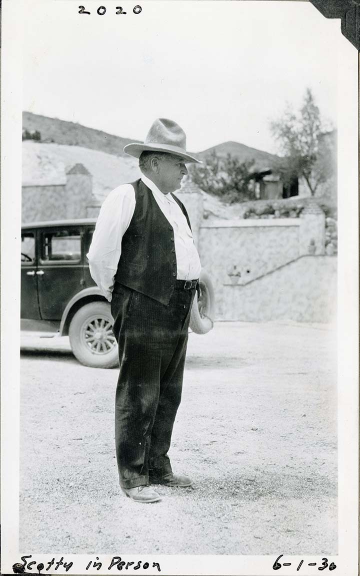 This is an historic black and white photograph from the Scotty's Castle Historic Photograph Collection, Death Valley National Park of older man standing profile with hands behind back wearing large cowboy hat, white shirt, and vest. Passenger car in background. Mottle stucco wall and landscaping out of focus in background. Inscription in black ink along upper and lower border.