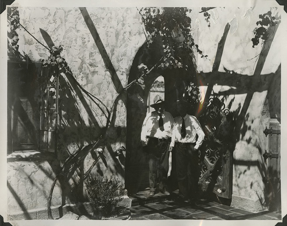 This is an historic black and white photograph from the Scotty's Castle Historic Photograph Collection, Death Valley National Park of Walter Edward Perry Scott and Albert M. Johnson posed by Scotty's Castle Main House and Annex small gate to Enclosed Patio. Circa 1930. Photographed by Mat Roy Thompson.