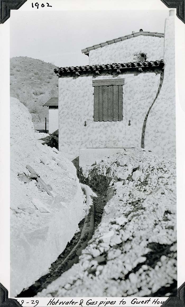This is an historic black and white photograph from the Scotty's Castle Historic Photograph Collection, Death Valley National Park of excavated trench in rocky soil with heavy pipe leading from foreground and curving left. Trench and pipeline disappear as it curves around behind a structure that has textured and two-tone stucco, Spanish tile roofline, textured wood shutters, and curve of exterior chimney flume. Inscriptions in black ink along upper and lower border.