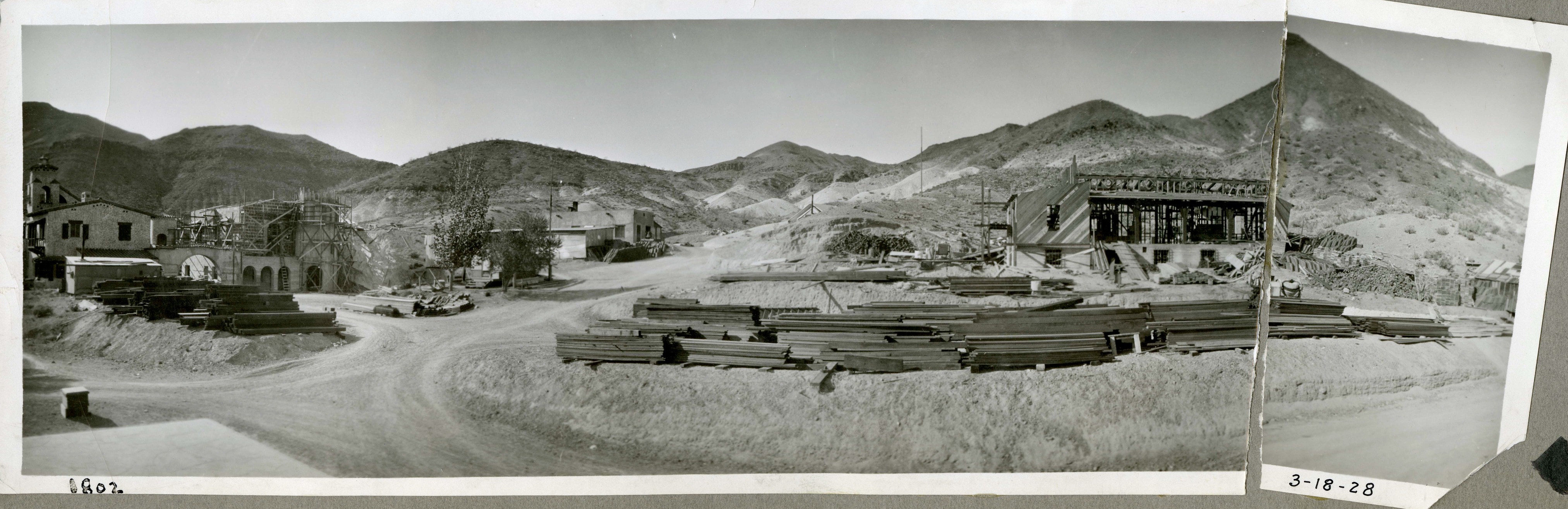 This is an historic black and white photograph from the Scotty's Castle Historic Photograph Collection, Death Valley National Park of a building complex. Large two-story building on each end of image under construction with scaffolding and forms. Extensive piles of lumber in front of buildings. Dirt, two-track roads lead in front of and between buildings. Barren mountains in background. Inscriptions in black ink along lower border.