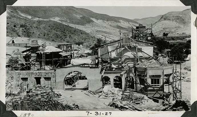 This is an historic black and white photograph from the Scotty's Castle Historic Photograph Collection, Death Valley National Park of Scotty's Castle Main House and Annex, looking east. Westside Enclosed Patio arched connecting wall constructed and stuccoed. Annex basement walls have hollow tiles in place. Annex roof sheathed. Main House Scotty's Bedroom Porch arched wall constructed. Canvas hanging over Main House walls. July 31, 1927. Photographed by Mat Roy Thompson.