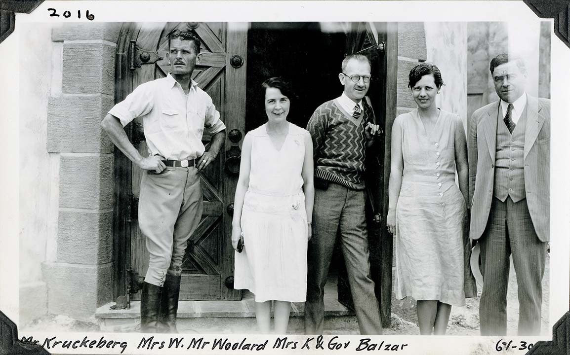 This is an historic black and white photograph from the Scotty's Castle Historic Photograph Collection, Death Valley National Park of two women standing between three men posing for camera in front of heavy wood-paneled door in arched opening. First man in white shirt, breeches, and calf-height leather boots. Second man in sweater with white shirt and tie, slacks, glasses. Third man in suit, tie, vest, and holding white hat. Both women in sleeveless dresses. Details of arched doorway and heavy wooden doors in background. Inscription in black ink along upper and lower border.