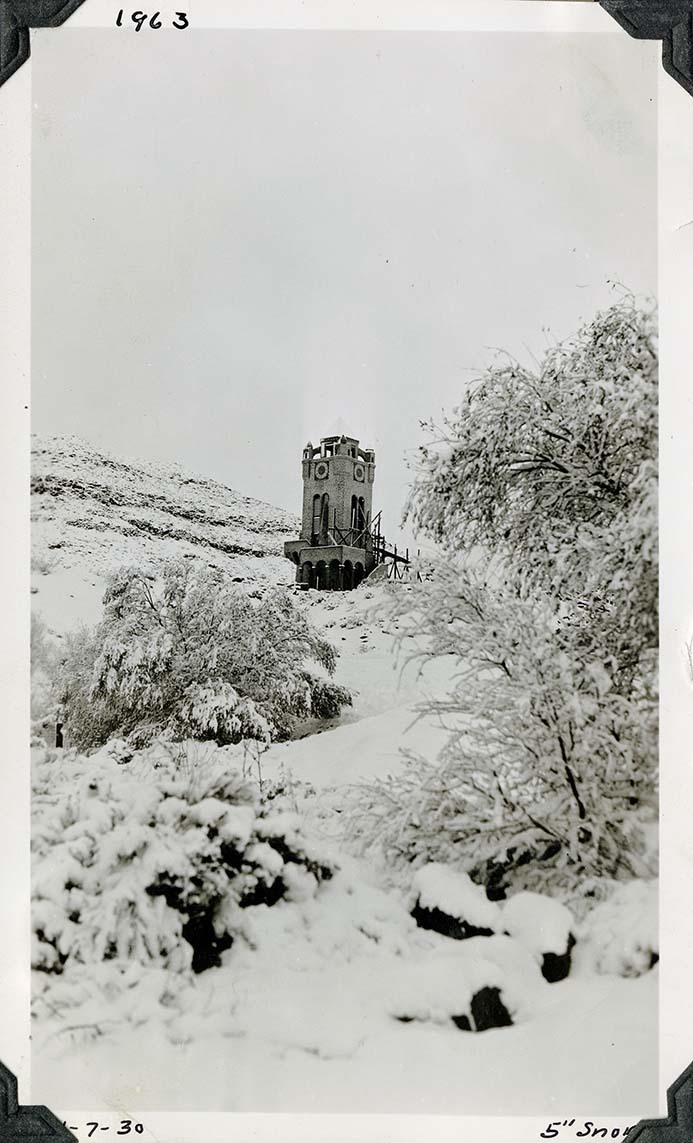This is an historic black and white photograph from the Scotty's Castle Historic Photograph Collection, Death Valley National Park of snow covered vegetation with tower in middle ground. Construction ramp leading to second story of tower. Inscriptions in black ink along upper and lower border.