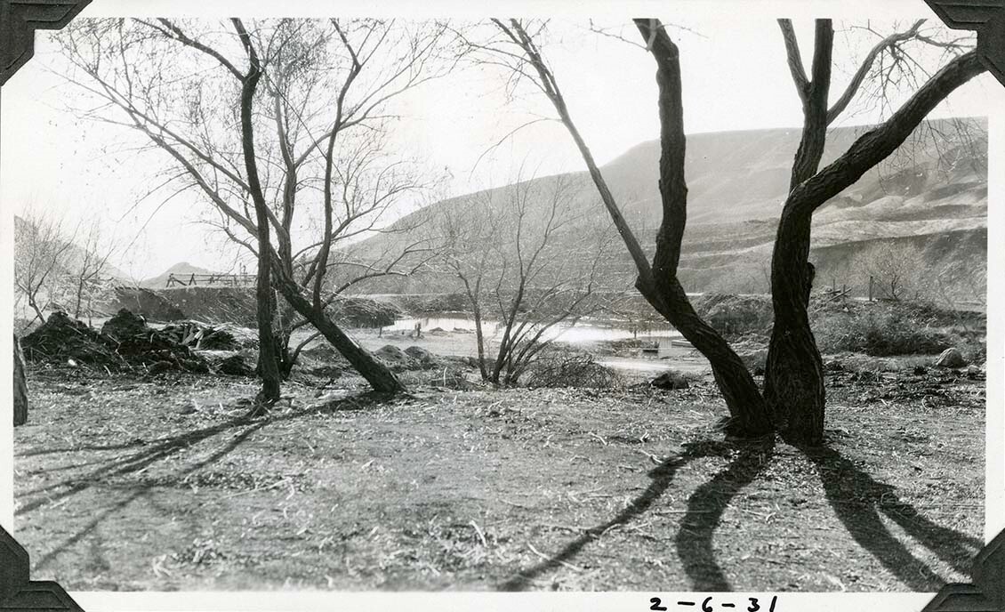 This is an historic black and white photograph from the Scotty's Castle Historic Photograph Collection, Death Valley National Park of mesquite-type tree trunks and branches and dirt in foreground. Piles of dirt and debris on left edge. In background is L-shaped dirt dam retaining water. Jack-leg type fence on top of dam on left. Dirt road between water and trees. Flat desert hillsides in background.