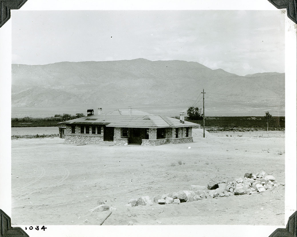 This is an historic black and white photograph from the Scotty's Castle Historic Photograph Collection, Death Valley National Park of newly constructed, square building. Rock stem walls, brick walls, hip roof with rolled roofing. Power pole to right. Pile of rocks in foreground. Field and distance mountains in background. Number in black ink in lower left corner.