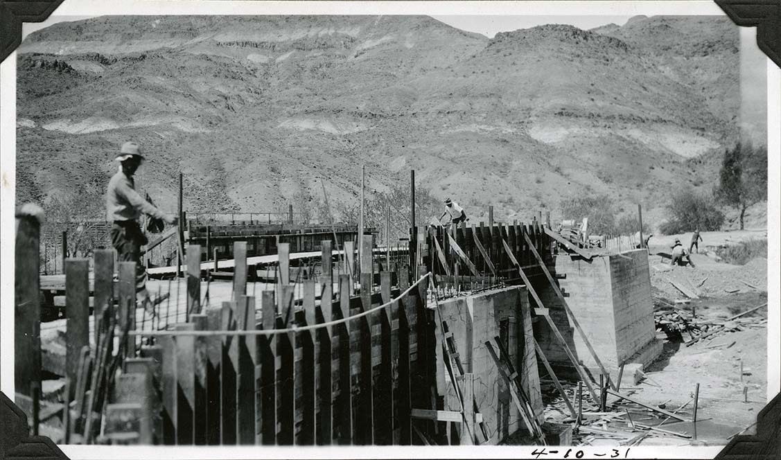 This is an historic black and white photograph from the Scotty's Castle Historic Photograph Collection, Death Valley National Park of simple concrete structure under construction. Two simple square structures complete. Elaborate wood forms in foreground for curved wall and decking between structures. Man working on forms in foreground. Another pushing concrete wheelbarrow over decking. At least four men in background shoveling sand pile. Large desert hillside in background.