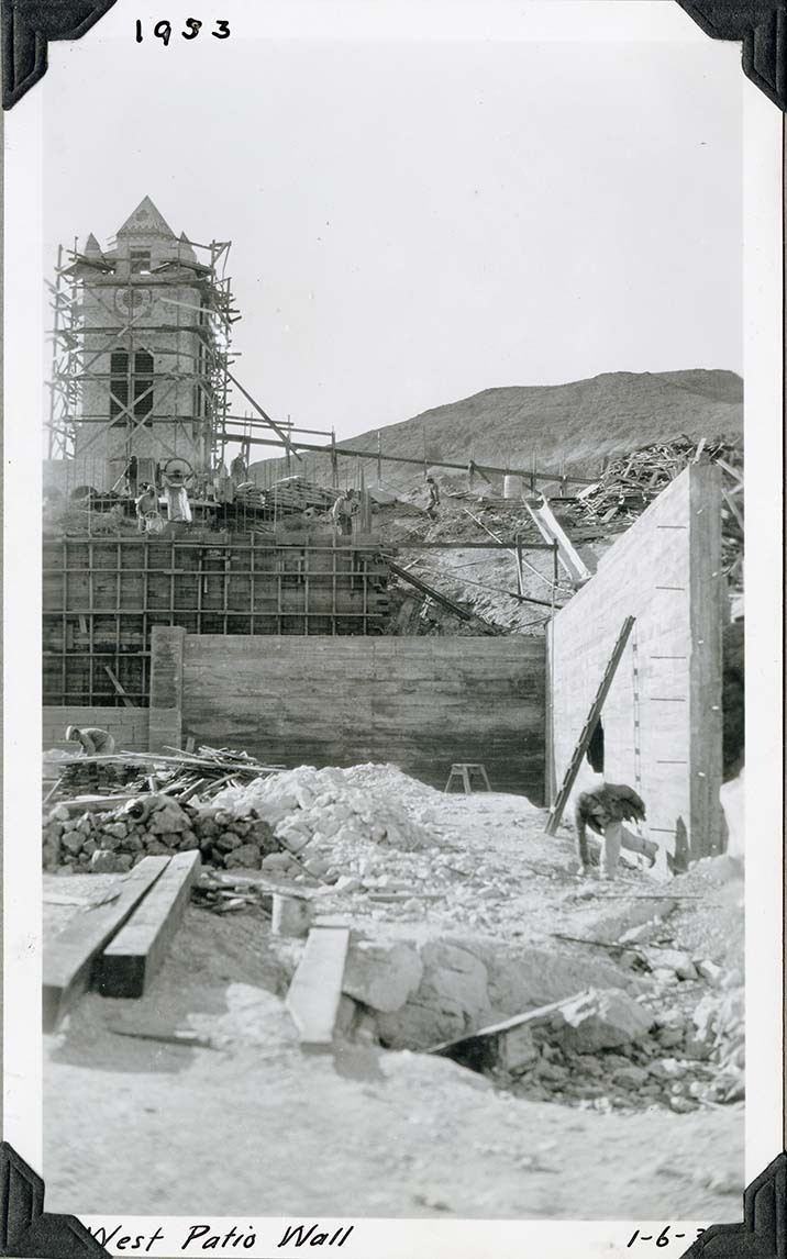 This is an historic black and white photograph from the Scotty's Castle Historic Photograph Collection, Death Valley National Park of busy construction site. Excavated rubble, lumber and worker in foreground. Completed concrete wall to left and middle ground with worker in front. Behind wall is wood forms for tall wall. Behind top of forms is concrete station with mixer, concrete barrow, workers, and bags of concrete. Behind this is a square tower covered with scaffold. Inscriptions in black ink along upper and lower border.