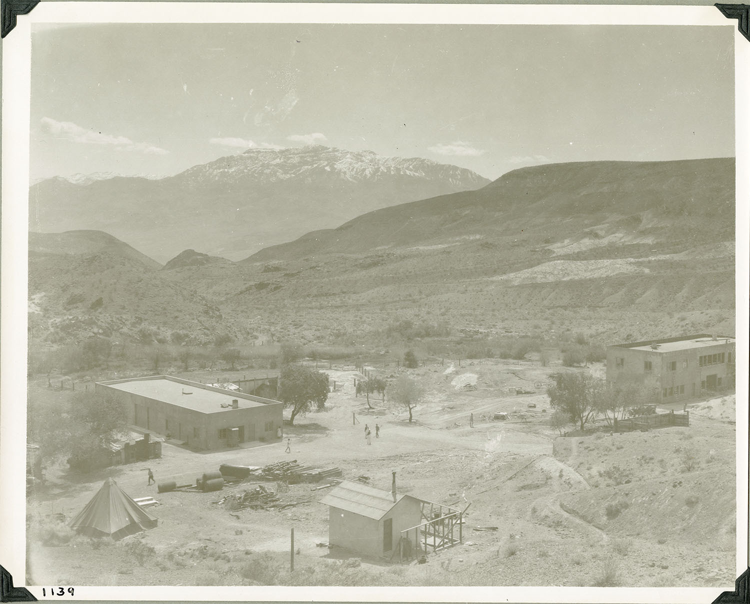 This is an historic black and white photograph from the Scotty's Castle Historic Photograph Collection, Death Valley National Park of a complex of simple structures in a valley surrounded by barren, desert hillsides. One tent structure and a very small, white building with railing around one side. A low, long rectangular building with a flat roof on left side. One opposite side a large, rectangular, two-story building with a flat roof. Dirt roads and some trees throughout complex. Number in black ink in lower left corner.