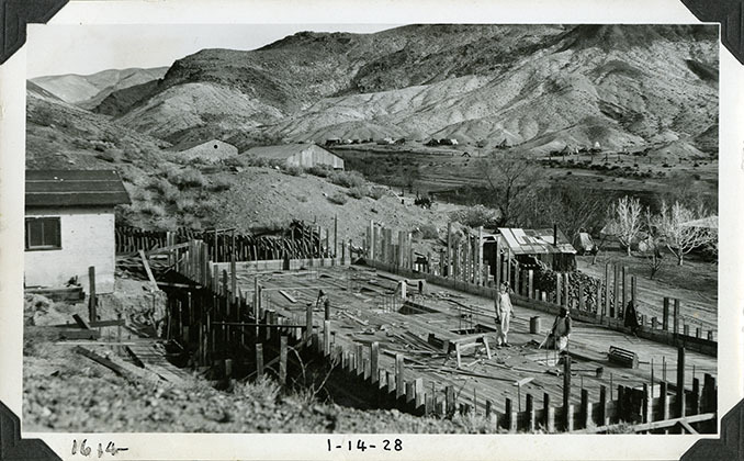 This is an historic black and white photograph from the Scotty's Castle Historic Photograph Collection, Death Valley National Park of Scotty's Castle Hacienda concrete basement and flat slab roof / floor poured. Two unidentified laborers. In distance, Indian Camp, Fire Cache, and Stables before remodeling. January 14, 1928. Photographed by Mat Roy Thompson.
