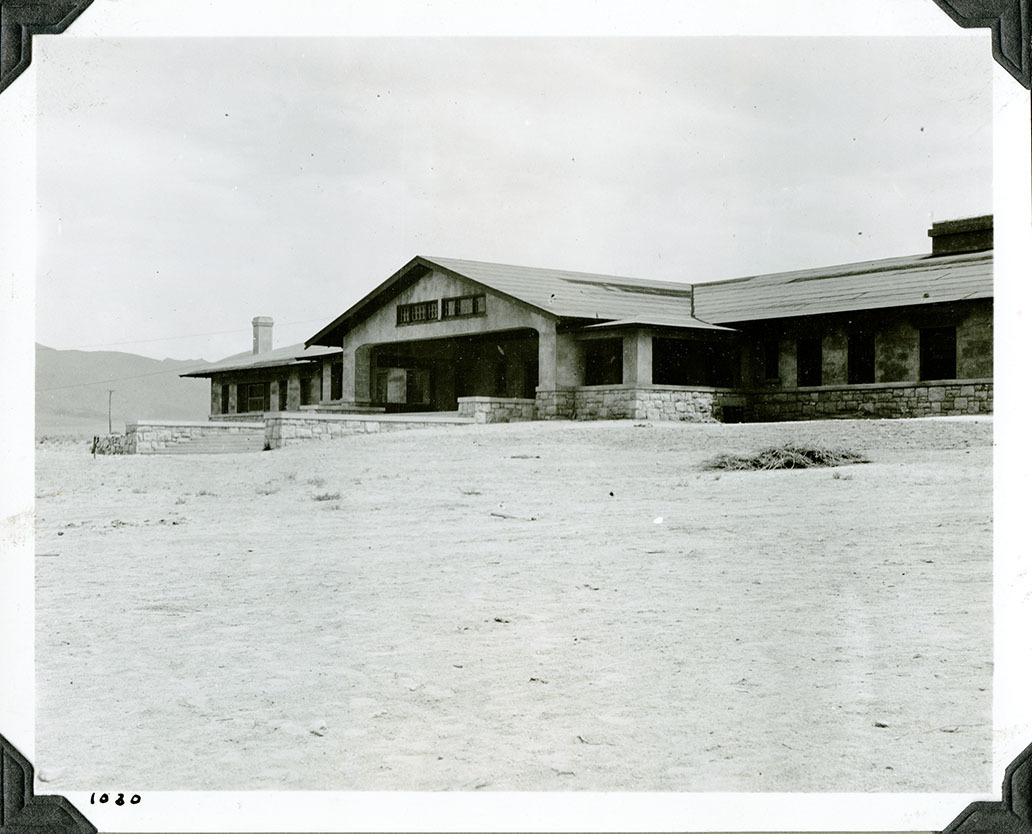 This is an historic black and white photograph from the Scotty's Castle Historic Photograph Collection, Death Valley National Park of large and long building newly constructed on dirt landscape. Rock stem walls, large covered front porch in center, long wings extend from central porch. Large windows along length. Low profile roof. Number in black ink in lower left corner.