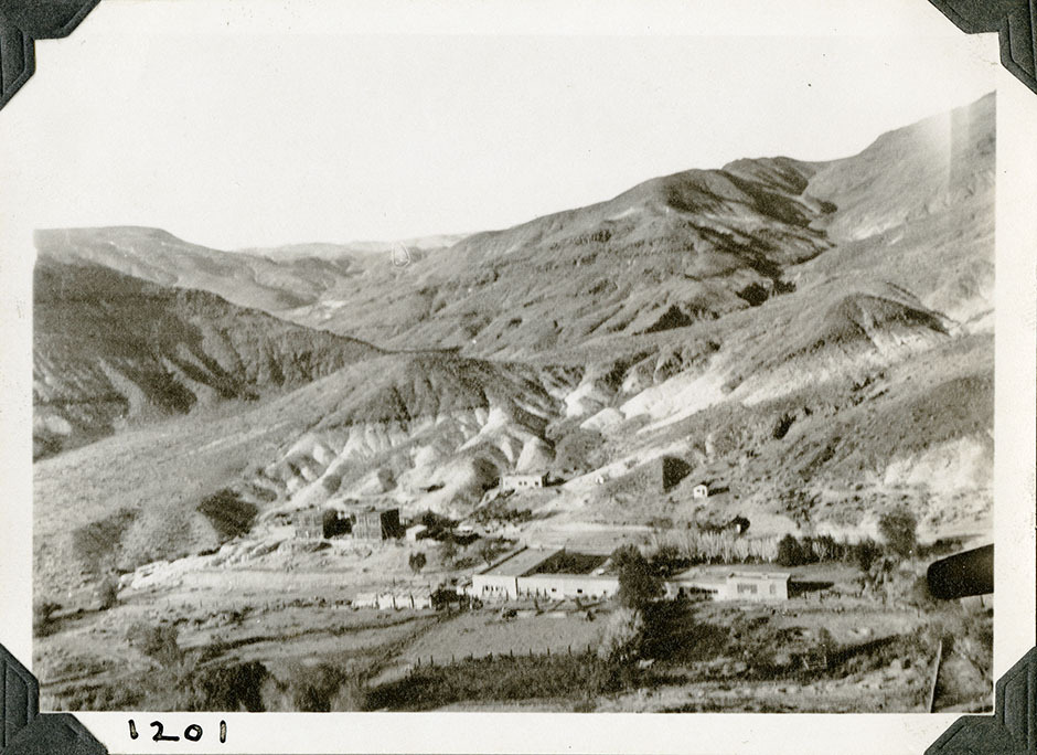 This is an historic black and white photograph from the Scotty's Castle Historic Photograph Collection, Death Valley National Park of a building complex within a desert landscape. One large structure under construction towards left. Large L-Shape building in center. Other small structures scattered across complex. Pastures and plantings defined by roads and fences. Upper half of image is looking across barren desert hills. Number in black ink in lower left corner.