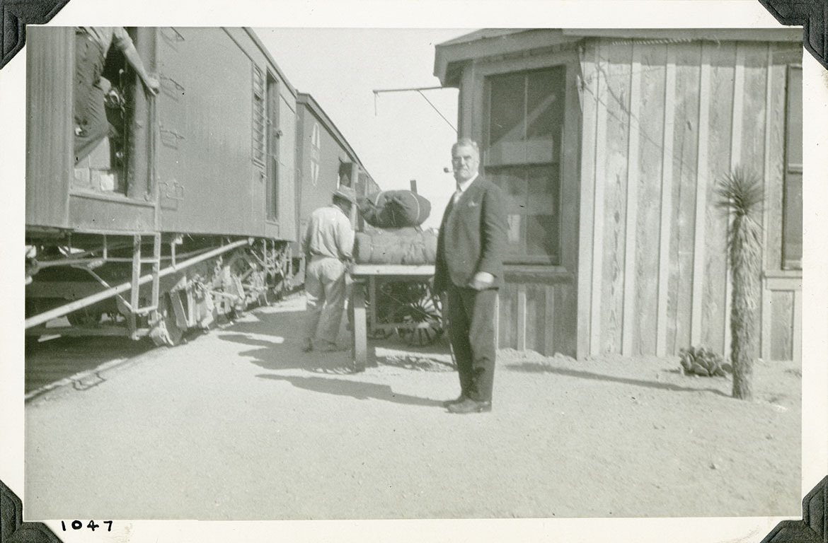 This is an historic black and white photograph from the Scotty's Castle Historic Photograph Collection, Death Valley National Park of main in suit standing in front of a delivery train on left, wagon in center, an board and bat building on right. Man standing in doorway of box car. Another man standing next to loaded wagon. Small Joshua tree on right. Number in black ink in lower left corner.