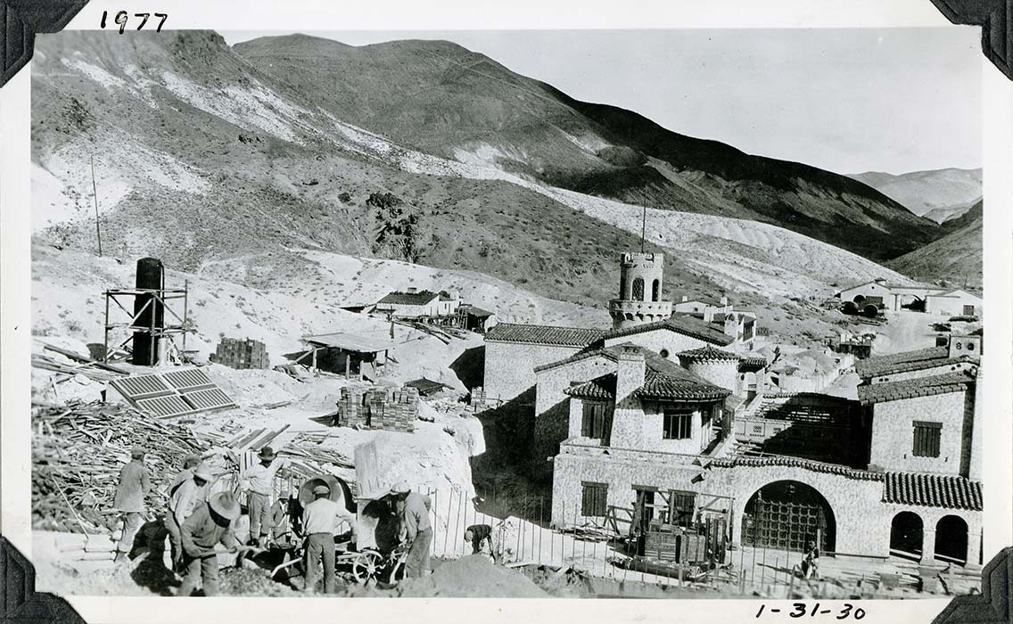 This is an historic black and white photograph from the Scotty's Castle Historic Photograph Collection, Death Valley National Park of at least nine men in left foreground working at mixing and pouring concrete. Overview of the rest of the building complex showing stages of completed remodeling and on-going construction.  Pile of lumber debris behind men. Solar panel and tall blank hot water tank to left. Man in distance at lower edge working in next to stacks of lumber and heavy-walled pipe. Look over roofline of Spanish style house – two sides connected by inner courtyard with arbor. Two workers in trench in distance in front of Spanish style building. Last building in distance two sections connect by courtyard gate. Several ranges of barren hills in background. Inscriptions in black ink along upper and lower border.
