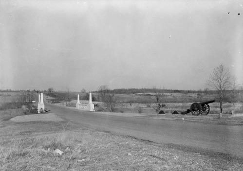 A narrow road passes past a cannon and over a bridge, lined by obelisks and ornamentation.