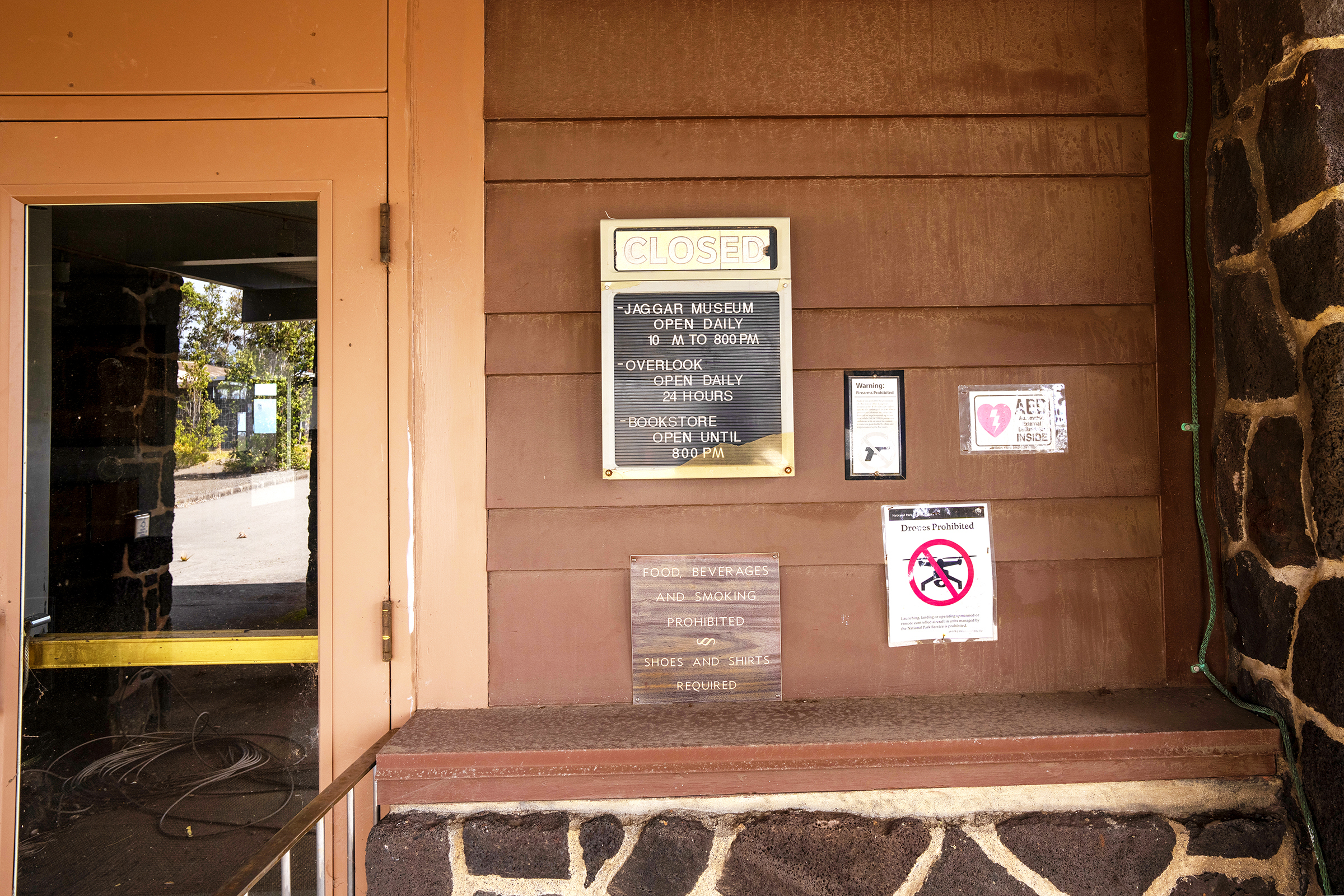 Several small signs outside a barricaded door. The prominent one reads CLOSED and has the Jaggar Museum hours. Antoher sign says No Drones 