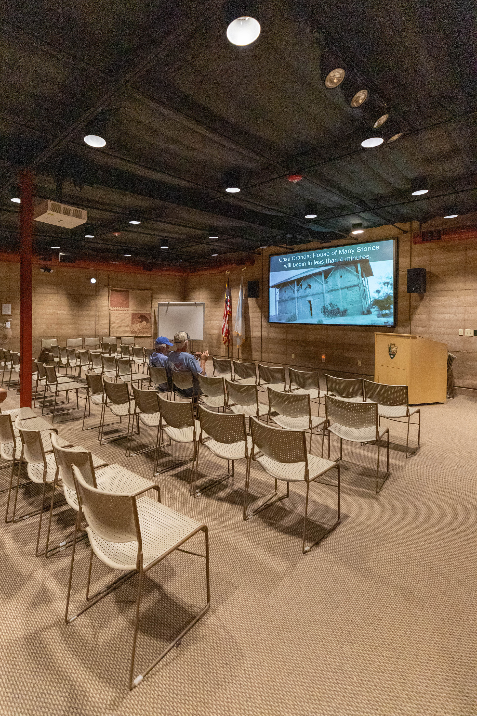 A room with a few dozen chairs facing a large screen. The screen has a photo on it which reads, Casa Grande: House of Many Stories will begin in less than 3 minutes. 