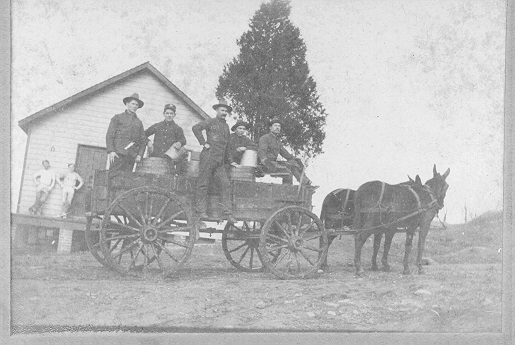 Two mules hauling a wagon full of barrels with 5 soldiers.  Wagon is in front of the bake house which has 2 men leaning against the wall.