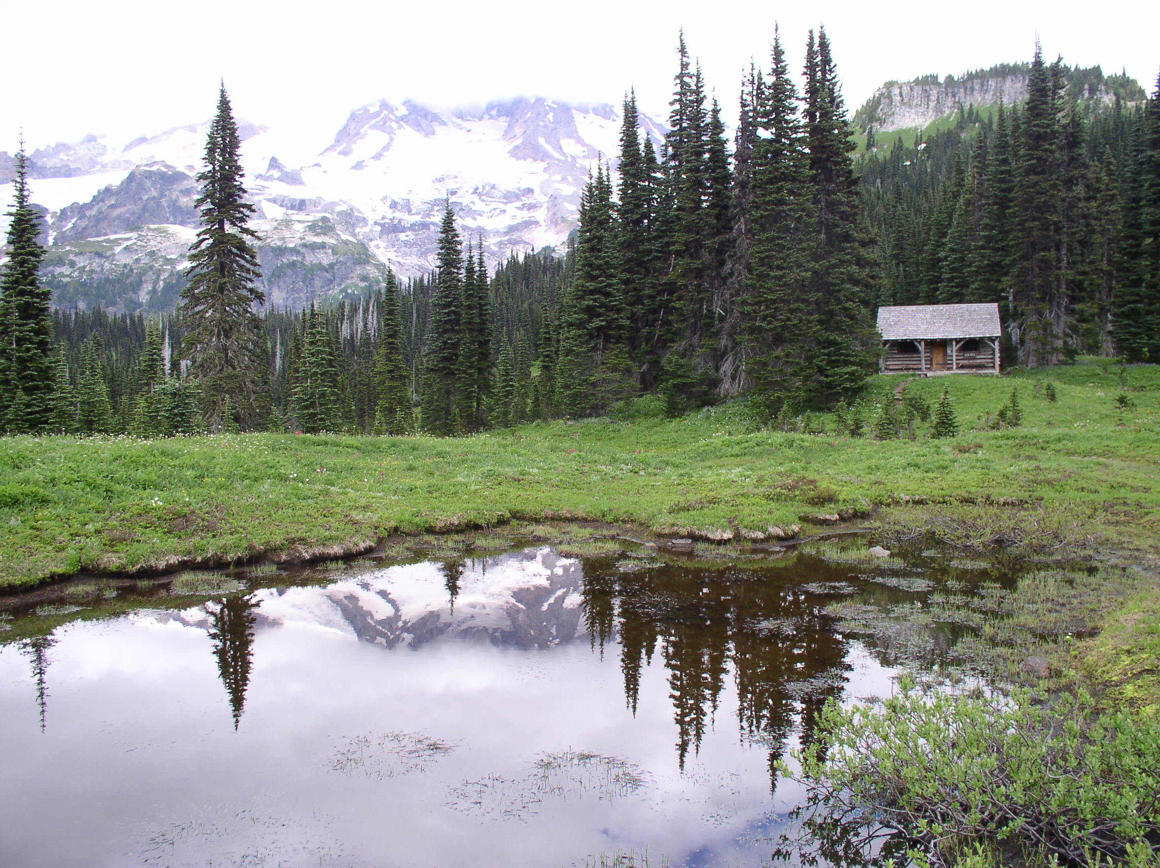 A still pond reflects a cloudy view of Mount Rainier in front of a small wood cabin tucked into the trees. 