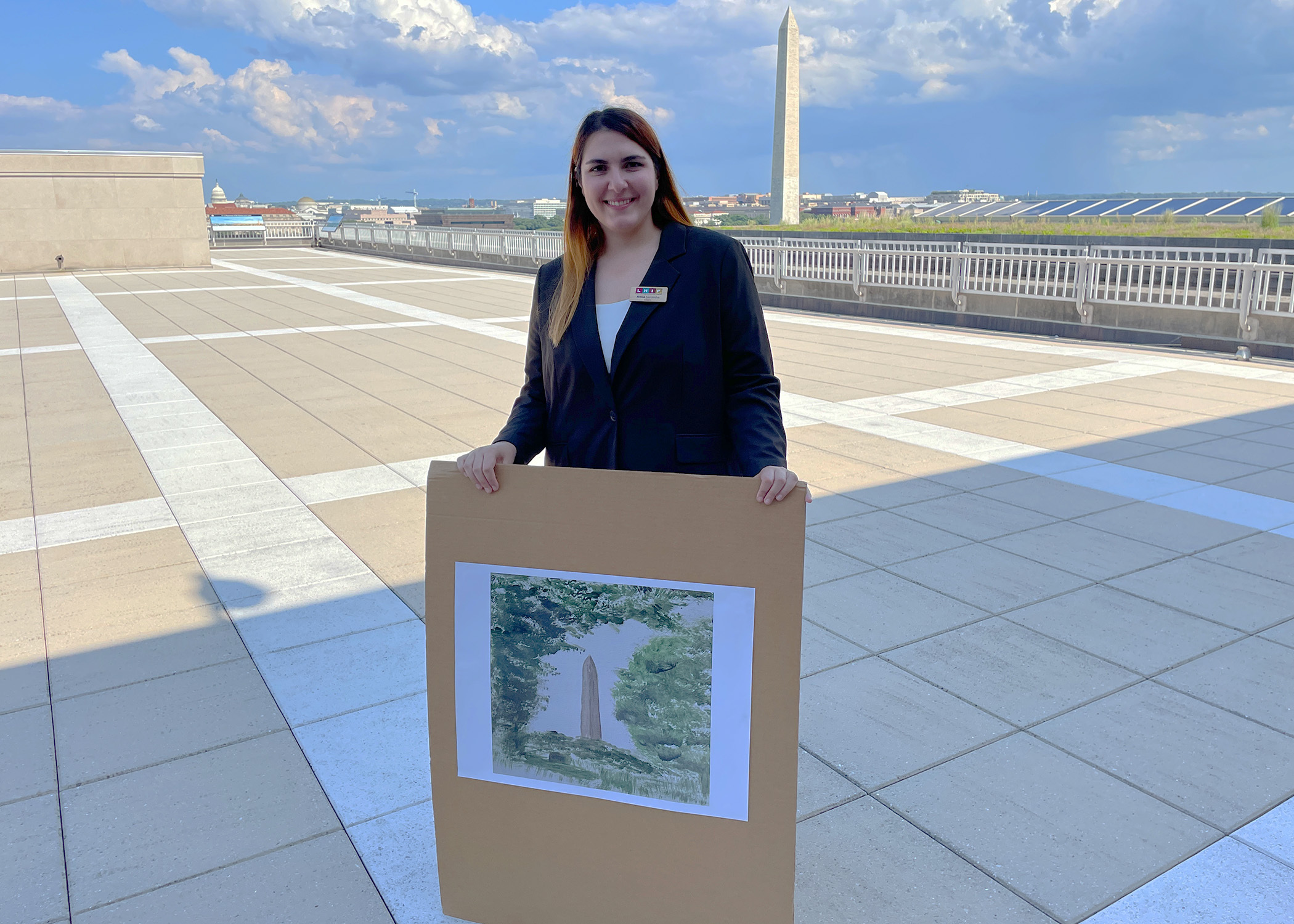 Person holding a tall cardboard with a picture of a painting of the Washington Monument. Person is standing at an open space with the Washington Monument in the background.
