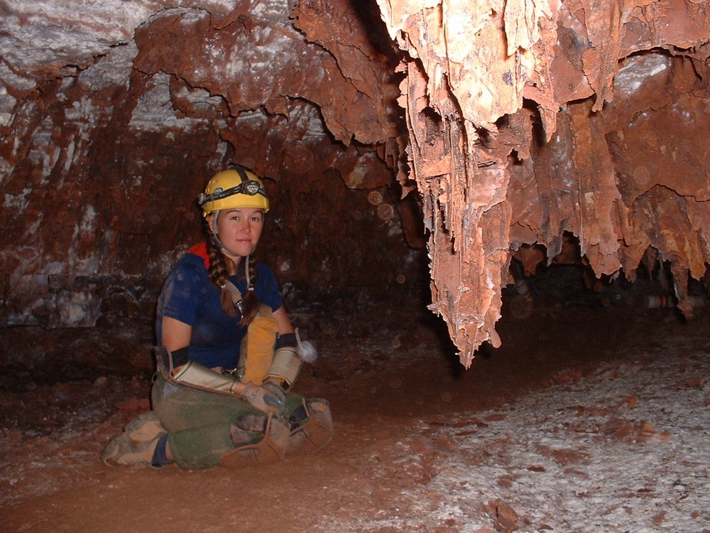  Long box shaped formations hang from the ceiling of a small cave passage. Under the formations, a woman is kneeling in her headlamp, knee pads, and elbow pads.