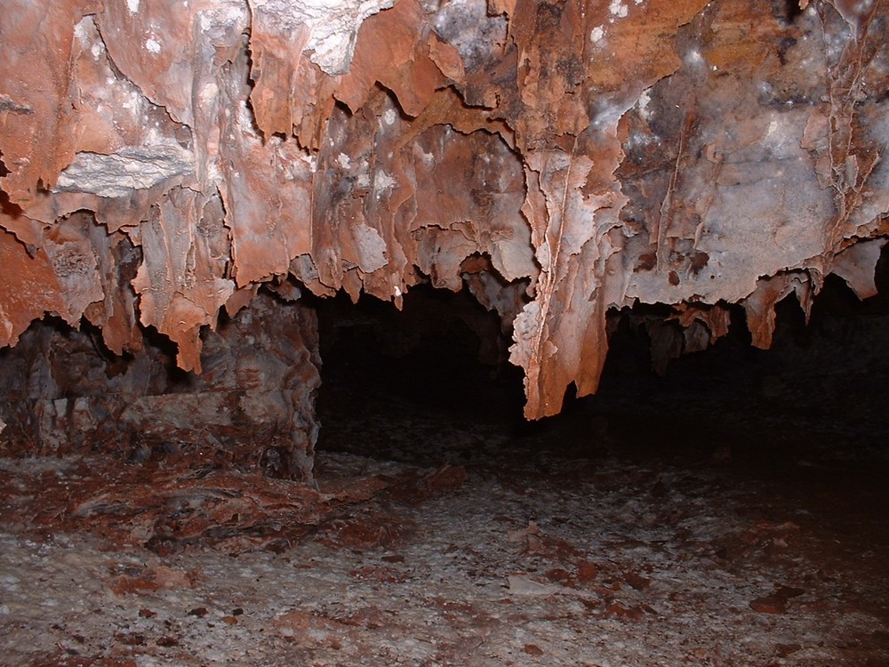 A long box shaped formation hangs from the ceiling of a small cave passage.