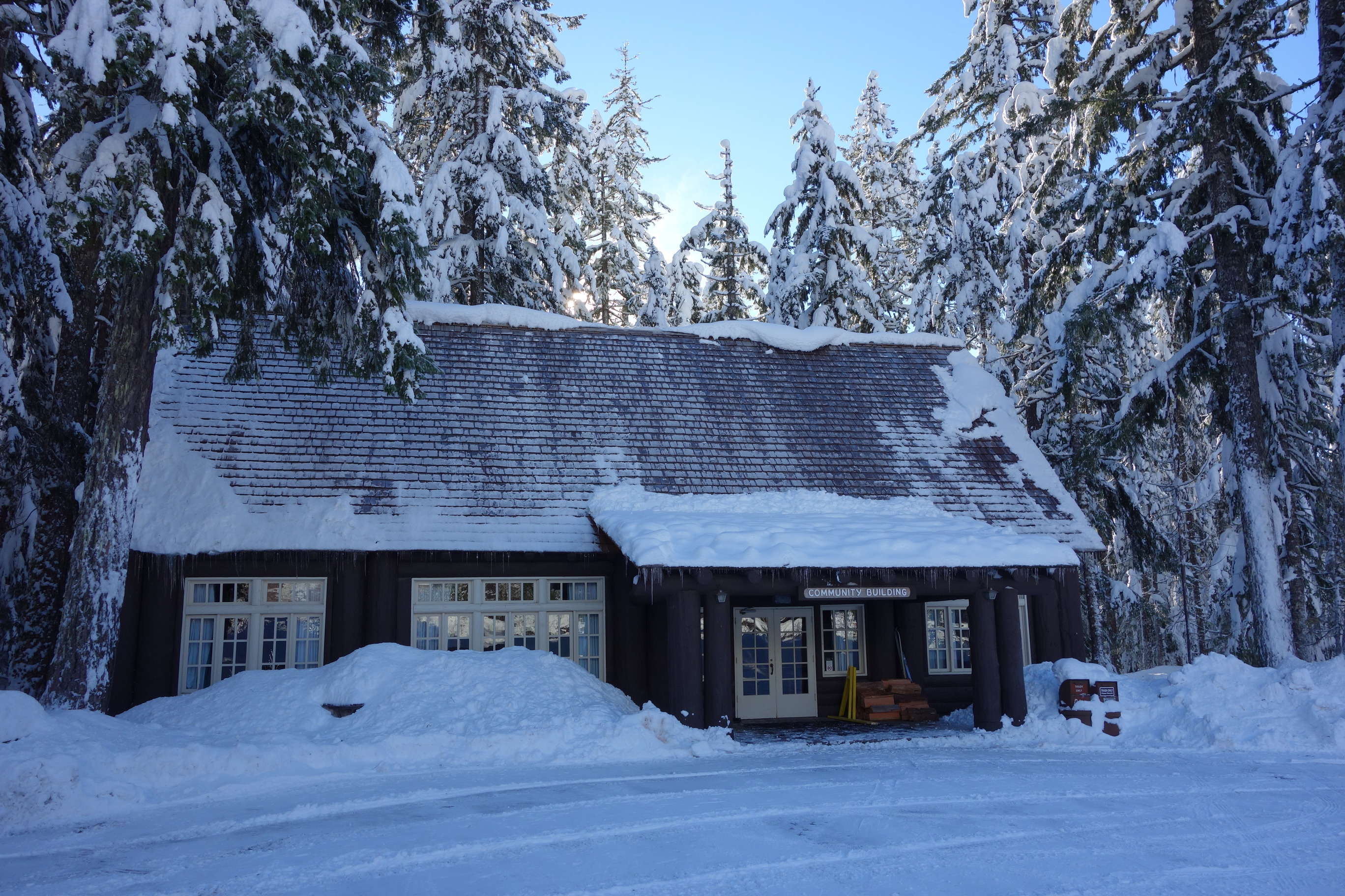 A large log building in a snowy forest with a steep shingle roof and a porch supported by log beams. The roof is covered in snow with icicles hanging off the porch. A sign over the porch reads “Community Building”. 