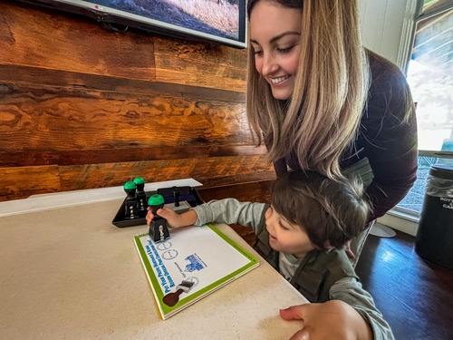 A child presses a stamp to a white page that has other stamps on it, and adult smiles down at the stamp book. 
