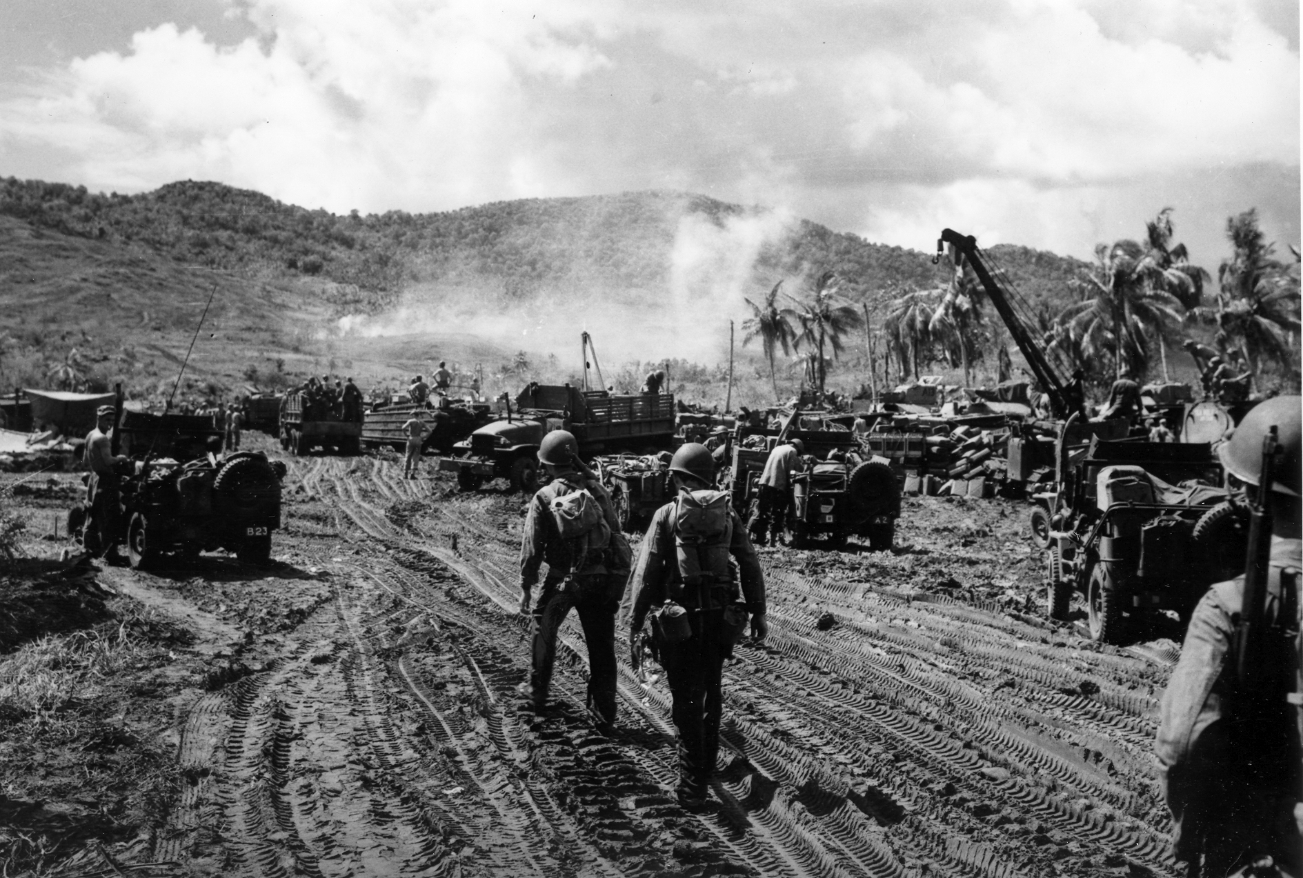Soldiers in helmets and gear navigate a muddy area with military vehicles and equipment, amidst a hilly, smoky landscape.