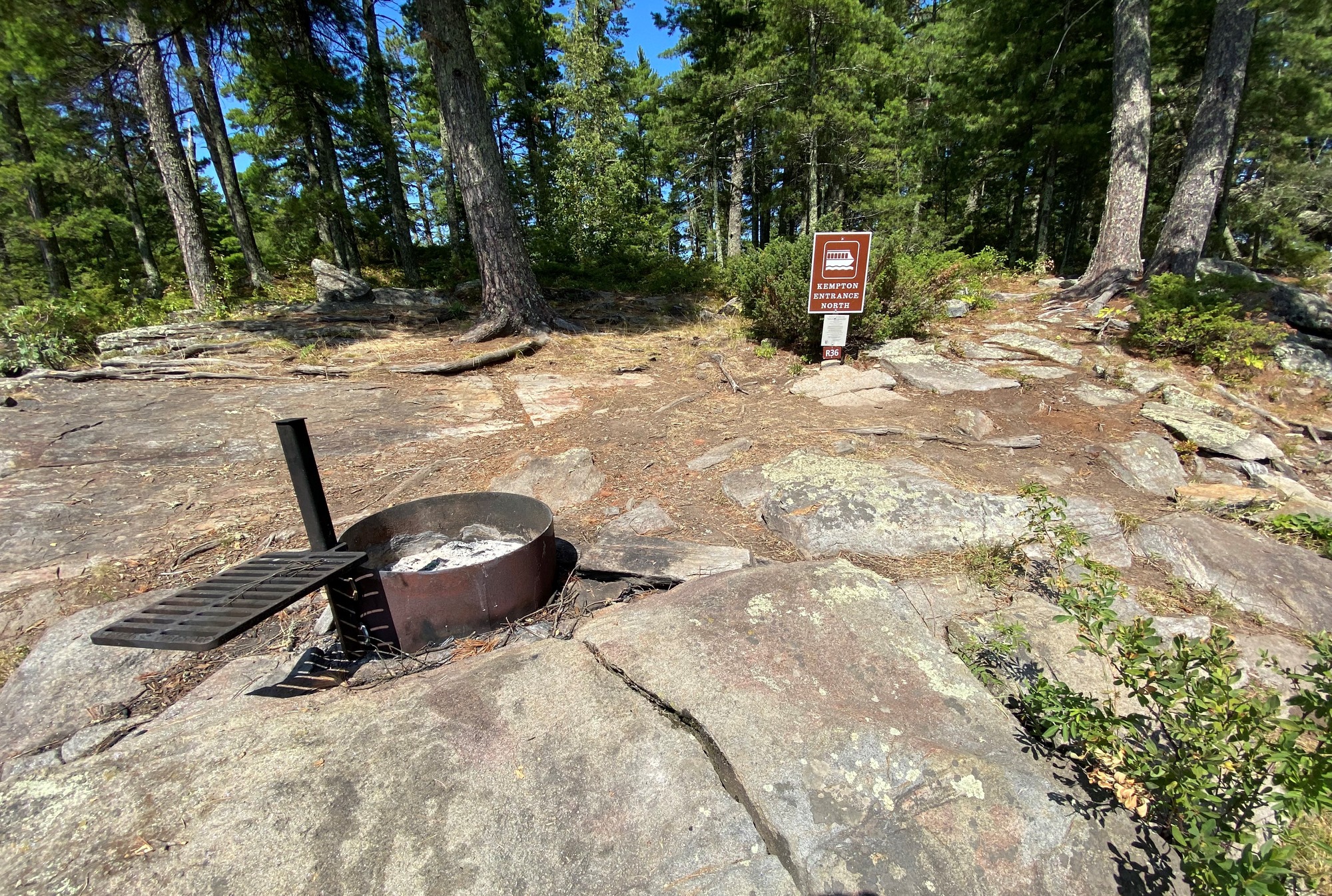 View of the houseboat site core of the houseboat sign and camp fire ring on rocky out cropping. Large pine trees are in the background surrounding the site.