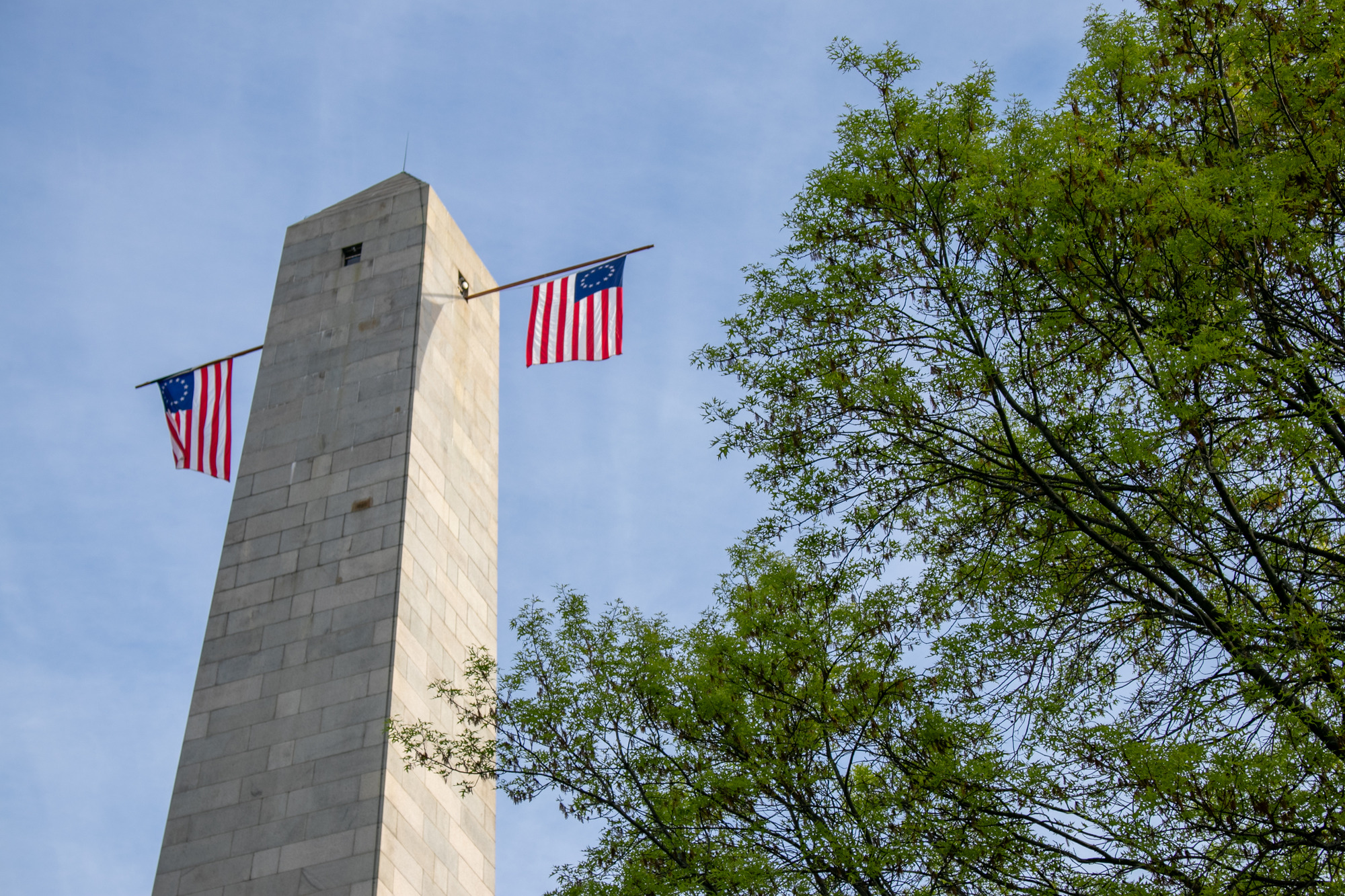 The top of a granite obelisk in morning sun, framed by green trees to the right. Two Betsy Ross US flags hang from opposite windows beneath the pyramidion.