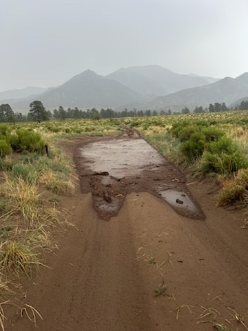 A dirt road with a large puddle with mountains and clouds in the background.