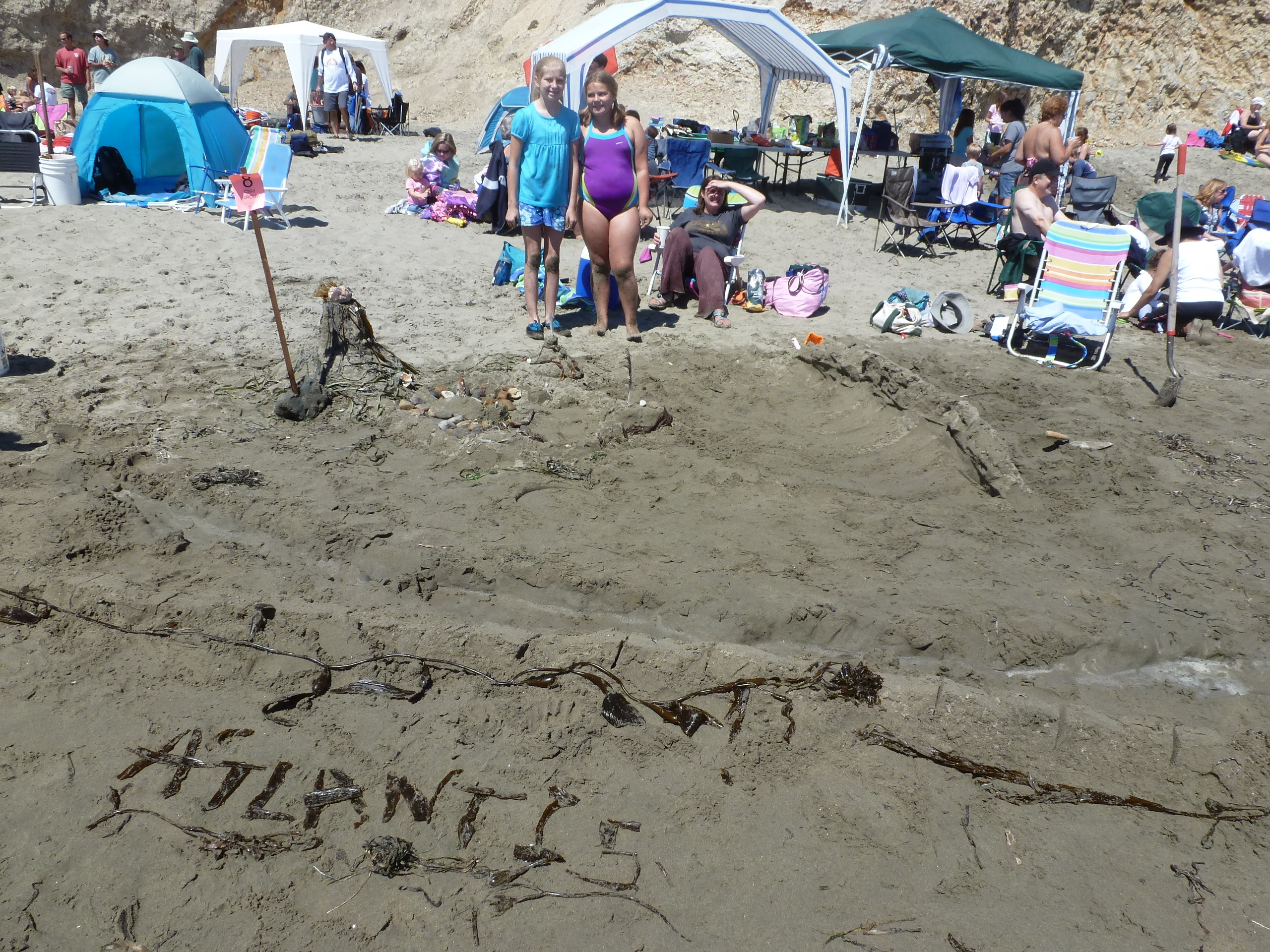Two girls stand behind a sand sculpture that is labeled Atlantis.
