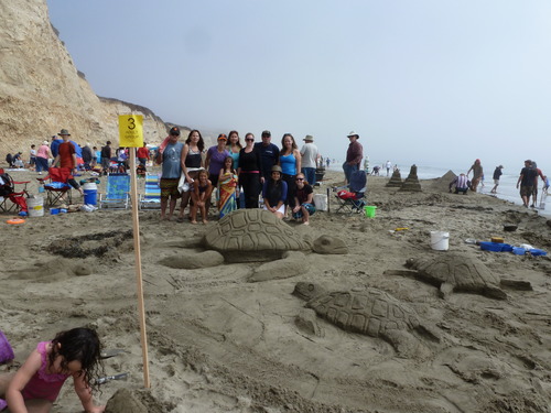 Eleven sand sculptors pose beyond a sand sculpture of a large sea turtle surrounded by three small sea turtles.