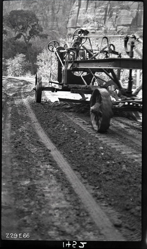Worker driving a road grader in Zion Canyon. Labeled 'experimental road paving. Processing sand and bitumul 60'.