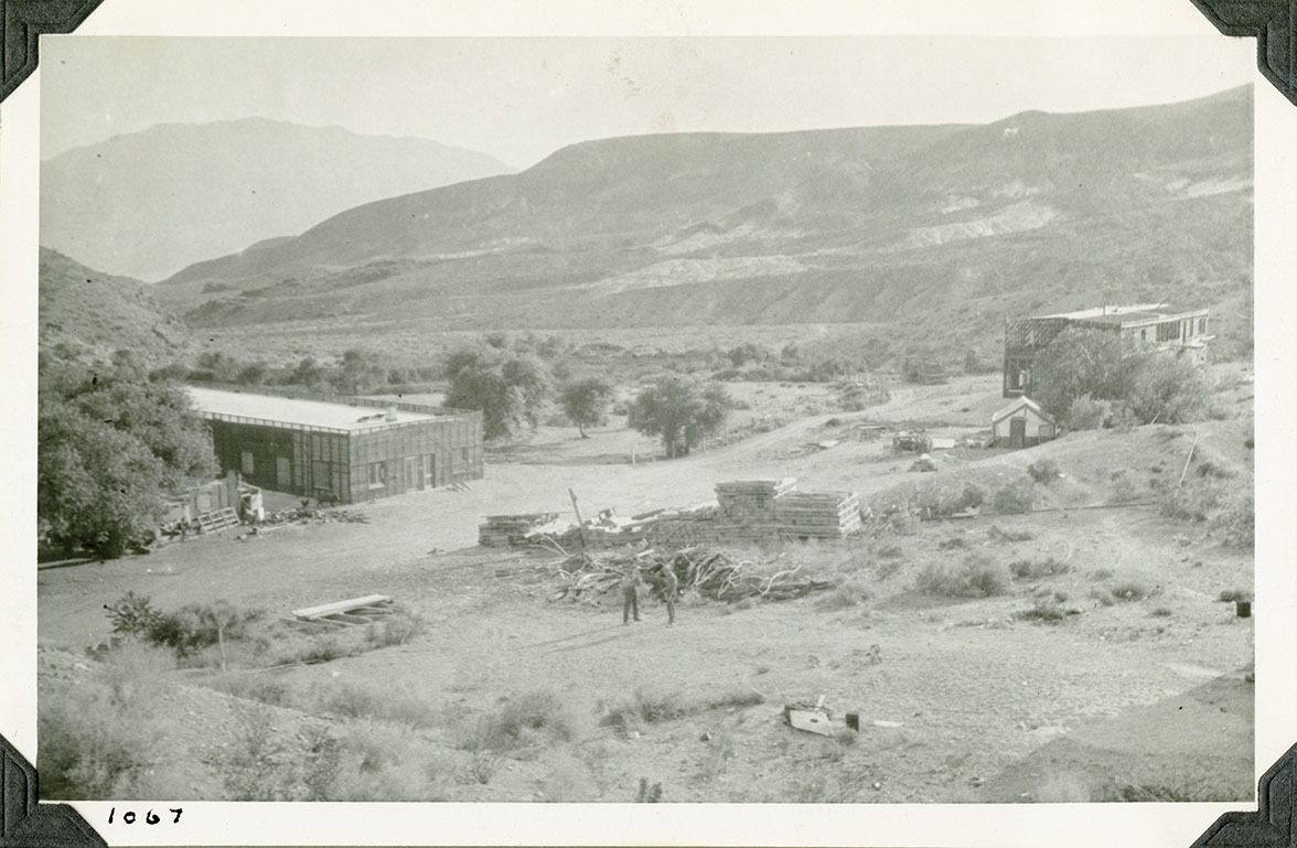 This is an historic black and white photograph from the Scotty's Castle Historic Photograph Collection, Death Valley National Park of construction in desert landscape. Large, two story, rectangular building in distance to right. Longer, single story, rectangular building to left. Construction material piles several locations. Roads leading through site. Two people towards center. Barren hillsides in background. Number in black ink in lower left corner.