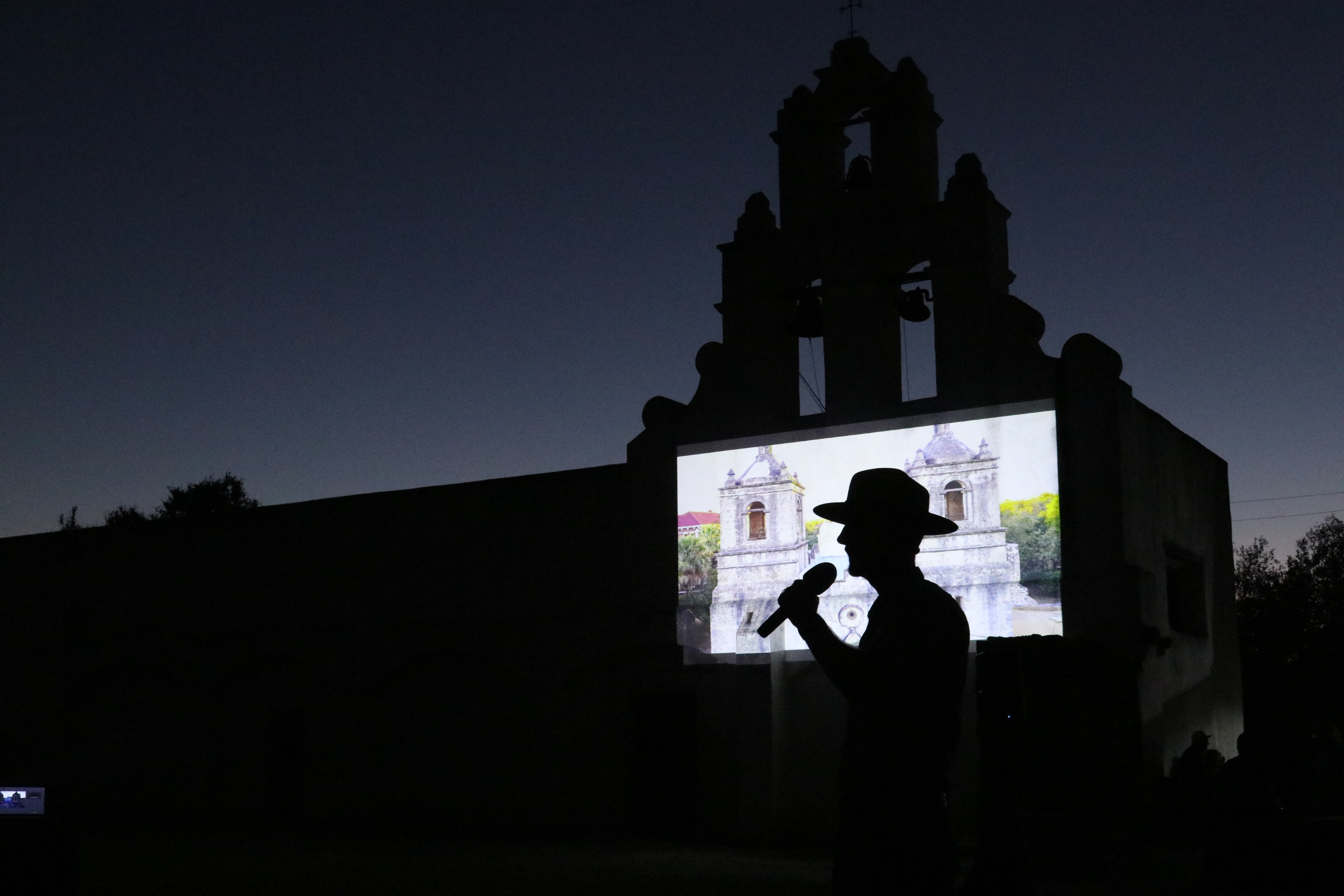 Park ranger speaking into a microphone at night with a projection behind him. 