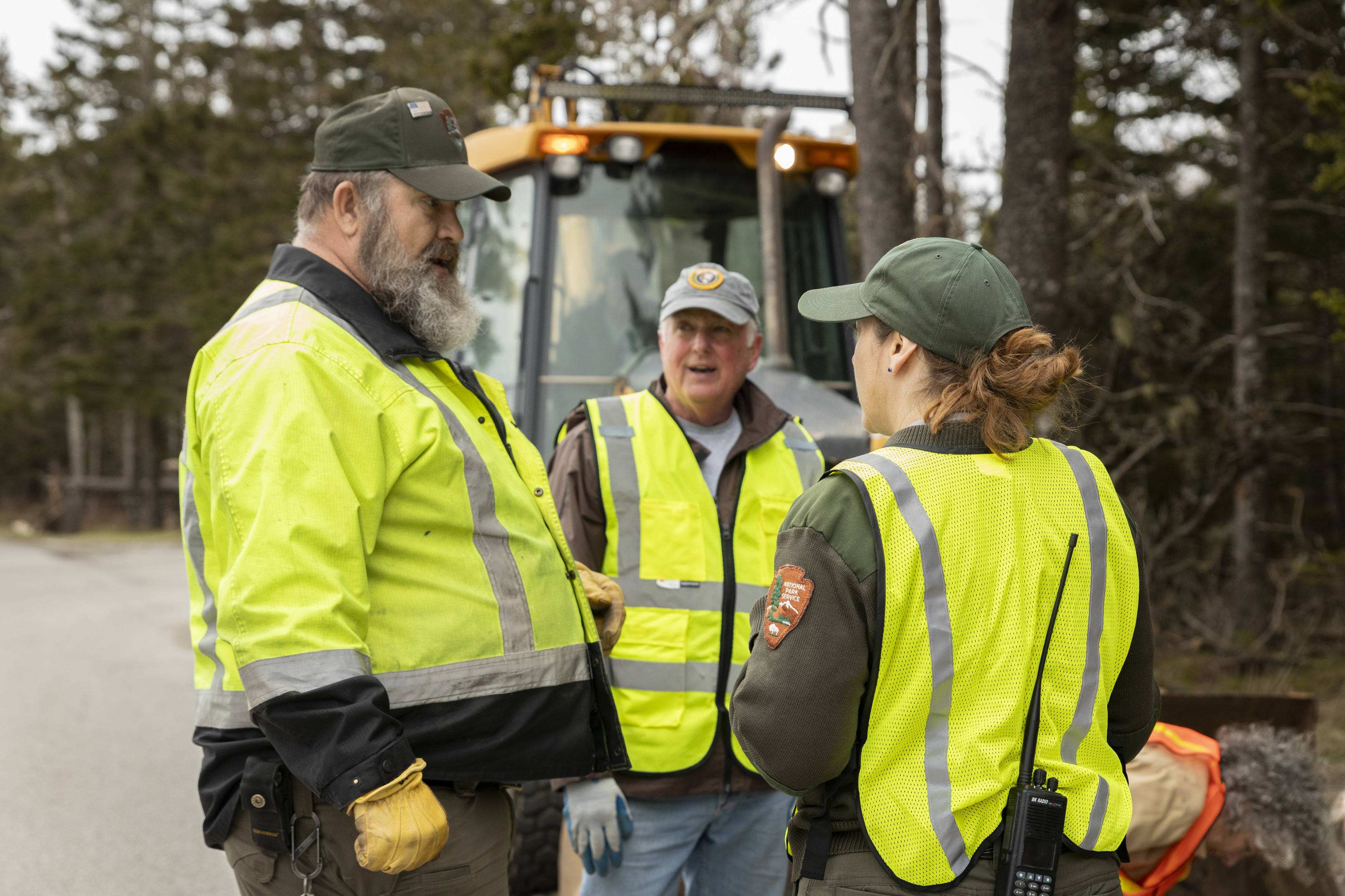 Three people in high-visibility vests, including two park rangers, having a discussion by the side of a road, with a forest in the background and a part of an backhoe visible.