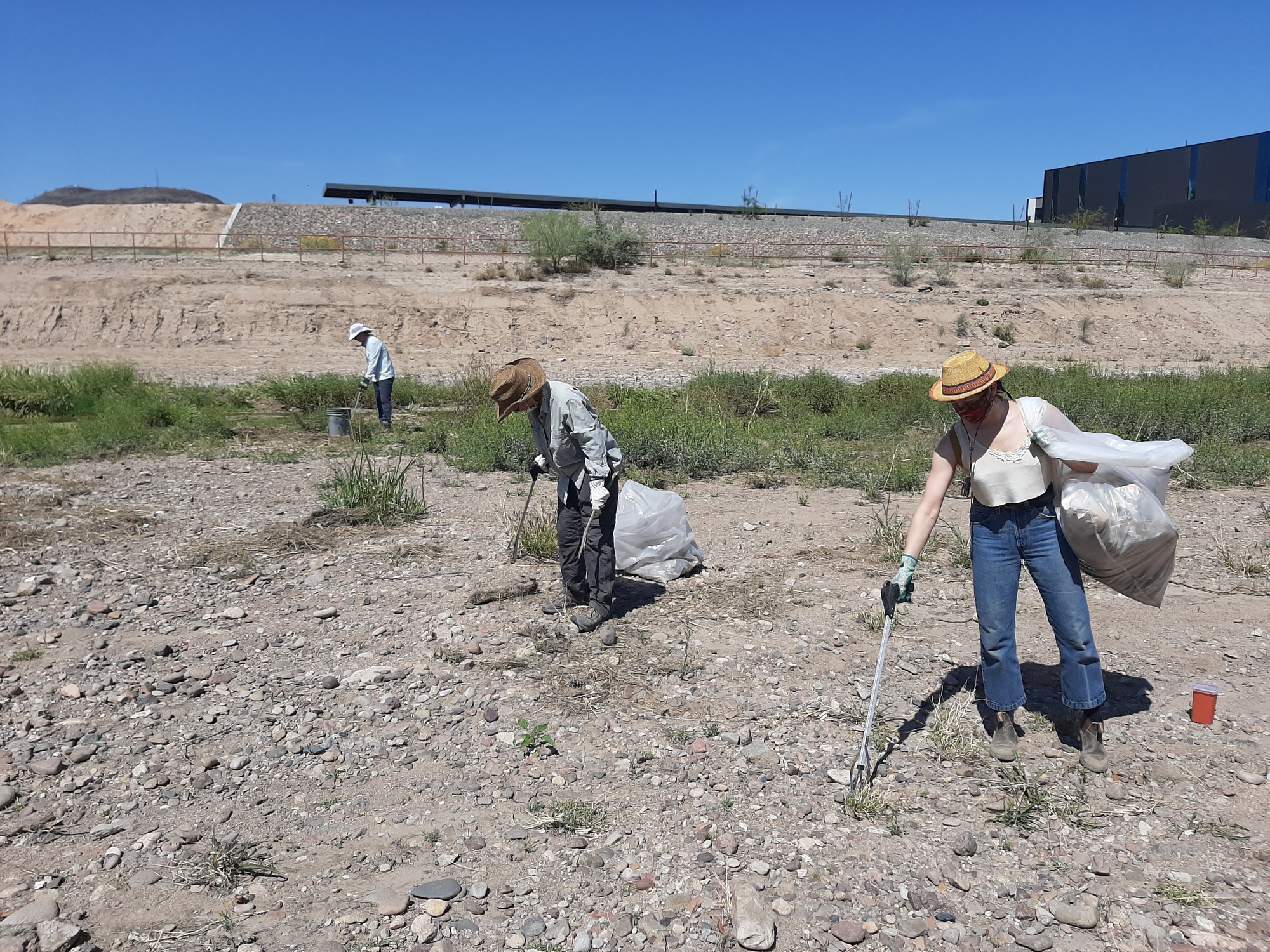 Three people wearing hats stand in a dry river bed picking up trash